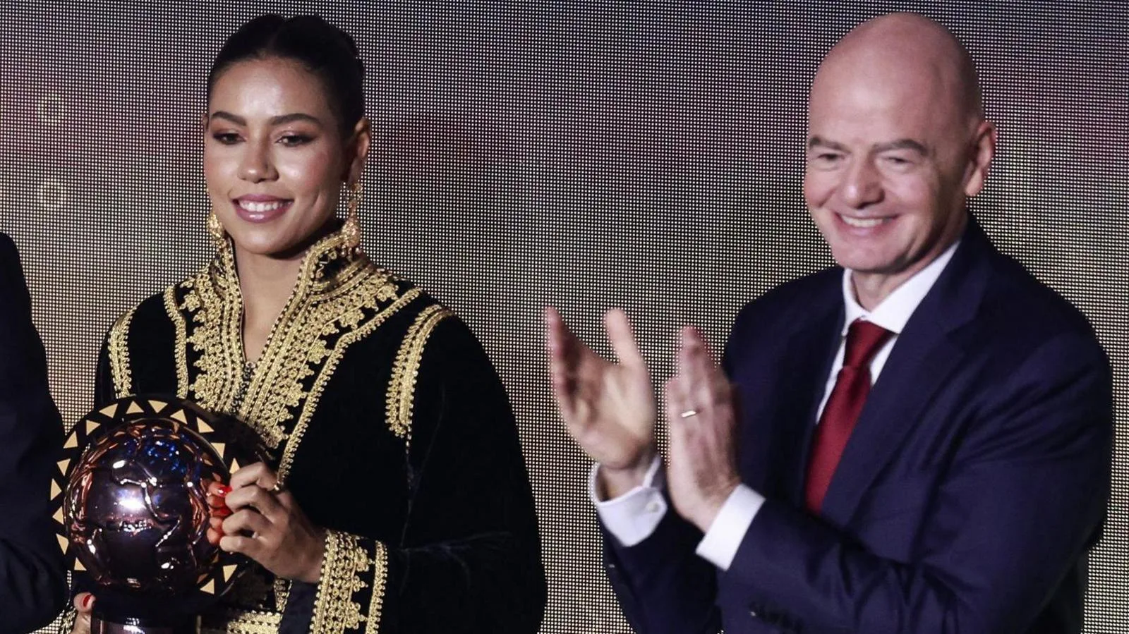 Ghizlane Chebbak smiles while wearing a traditional style black dress with elaborate golden detail and holding the Caf Women's Footballer of the Year award, which features a shining metallic football positioned inside a round thin round perimeter that runs around the vertical circumference. She stands next to Fifa president Gianni Infantino, who is wearing a blue suit, white shirt and red tie while smiling and applauding. Both are visible from the waist up