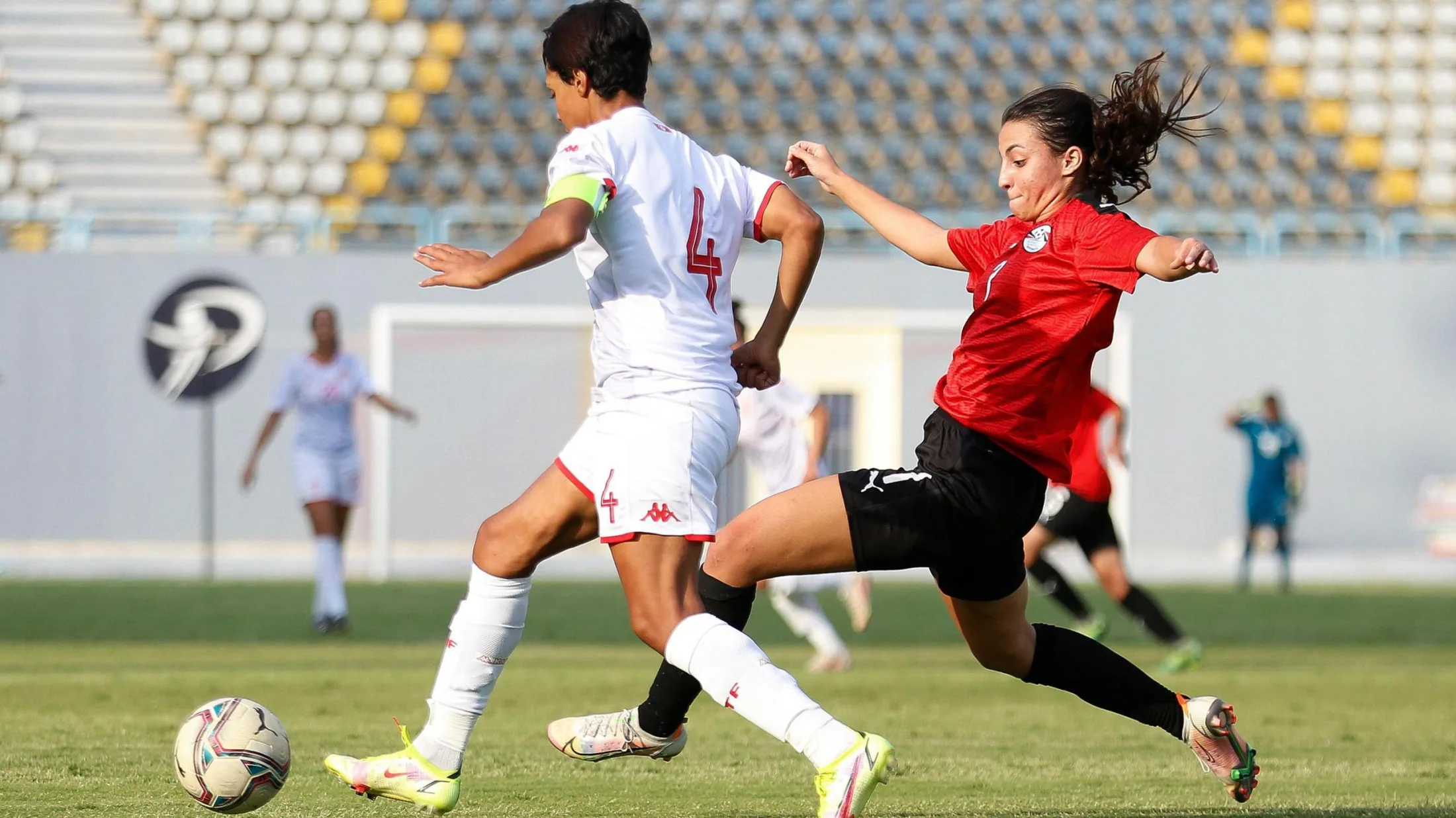 An action shot during a football match shows Nadine Ghazi, wearing a red Egypt shirt, black shorts, black socks and white boots, spreading her arms for balance and stretching her left leg forward as she challenges an opponent in an all-white strip for the ball. Four other players, a goal and an empty stand can all be seen out of focus in the background.