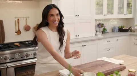 Meghan calls Harry 'a charmer' during MasterChef Australia appearance Netflix The Duchess of Sussex in a kitchen smiling and wearing a white outfit