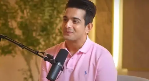 Beerbiceps via YouTube Ranveer Allahbadia during his podcast. He is wearing a light pink polo shirt and is sitting in front of a mic in a warmly lit studio with yellow interiors. 