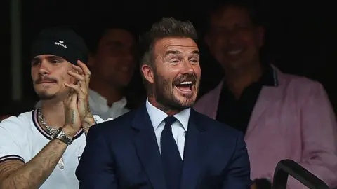 Getty Images English former football player David Beckham smiles during the 2026 World Cup football tournament quarter-final match between Norway and England at Miami Stadium in Miami Gardens on July 11. He is wearing a blue suit, white shirt and a blue tie.