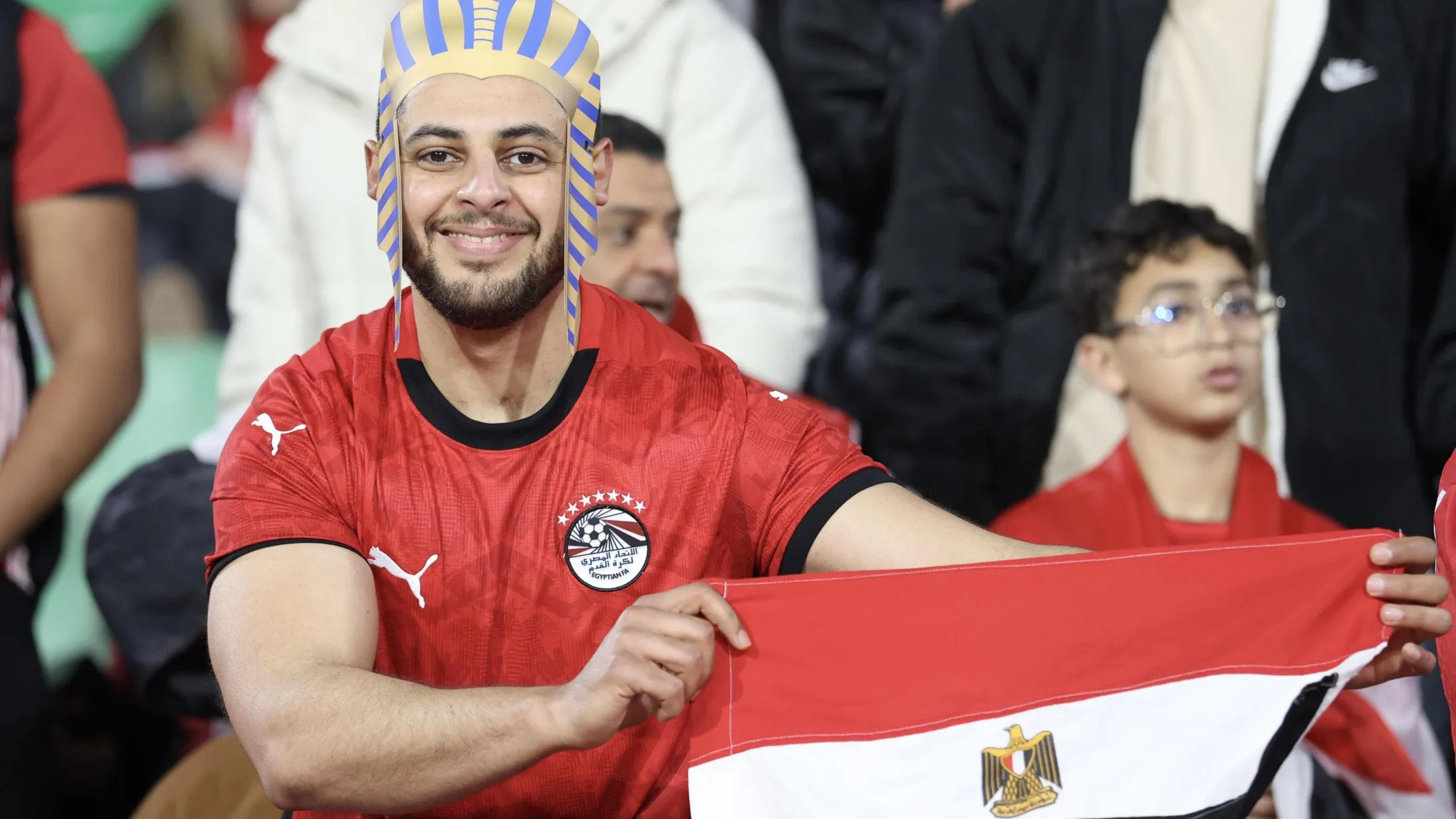 A smiling Egypt fan visible from waist up wears a red national team football shirt and a Pharaoh-style headdress as he holds up an Egyptian flag. Other fans can be seen out of focus in the background