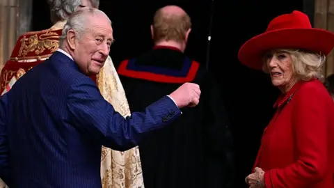 AFP via Getty Images The King and Queen turn around to look behind them. The King holds a hand up while the Queen smiles