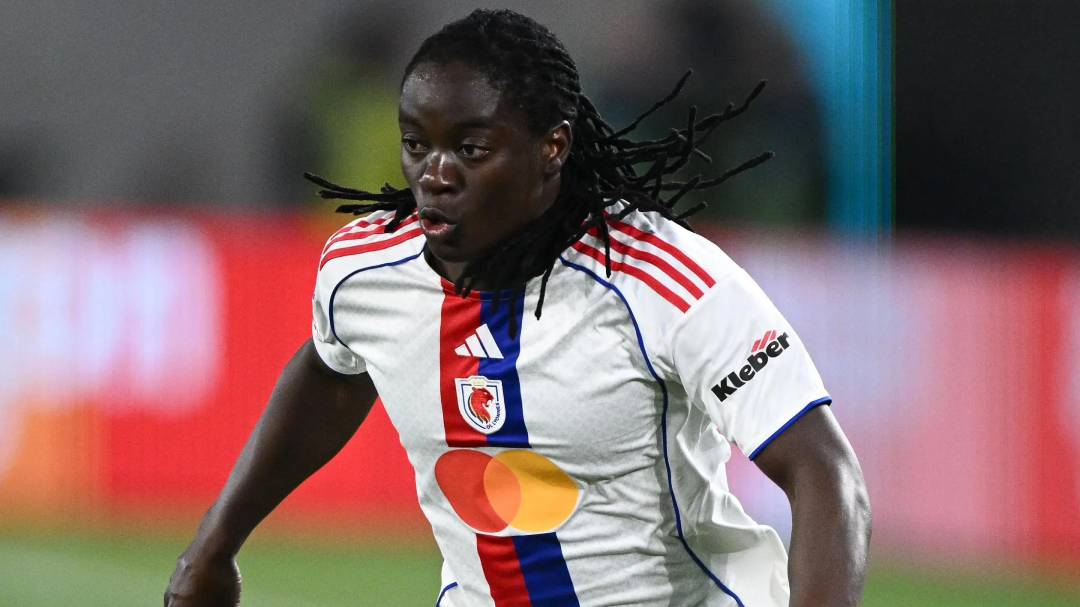 Tabitha Chawinga visible from waist up while in action wearing a white OL Lyonnes shirt with red and blue detail running down its centre. She puffs out her cheeks as she runs during a football match, with her many braids flapping in all directions behind and to the side of her head
