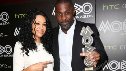 PA Media Kanya King and Idris Elba standing together, she's pointing at him and he's holding a Mobo trophy