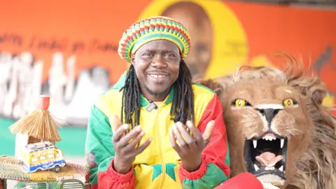 Aliou Ngom A man wearing a colourful yellow, red and green top with hat in matching colours looks at the camera. He has long hair. There is a large replica of a lion's head on the right of the picture with bared teeth and yellow eyes. 