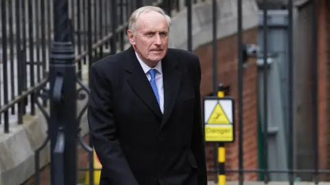 PA Media Paul Dacre, wearing a black coat, white shirt and blue tie, arrives at the Royal Courts Of Justice