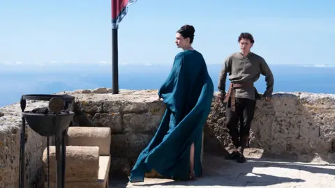 Universal Pictures Anne Hathaway as Penelope (Odysseus's wife) in centre of image in blue gown looking to the side and Tom Holland as their son Telemachus leaning on wall in khaki top and dark trousers.