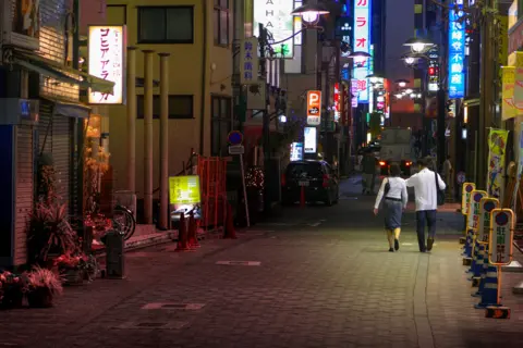 Getty Images A couple, a woman and man, walk in a Toyko street at night.  They are both wearing white shirts and grey bottoms and illuminated signs are seen in the backgorund.