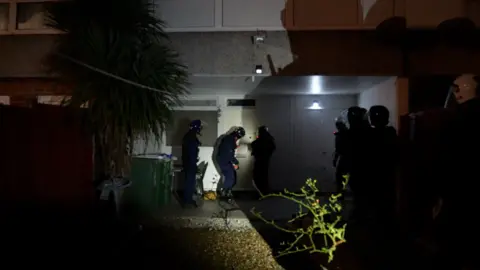 Dozens of romance scammers arrested after specialist fraud squad investigation in UK, Nigeria Three police officers outside a ground floor apartment at night time, dressed in helmets and riot gear, preparing to knock down the door
