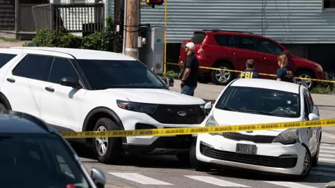 Getty Images Two white cars that collided are behind yellow police tape. The windscreen of one car has bullet holes in it