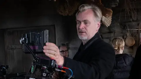 Universal Pictures Christopher Nolan in the centre of the photo and is looking through an Imax camera. He is wearing a black jacket.