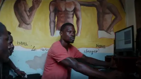 The notorious Cameroonian prison with its own record label Artists Equity Rapper Stone uses a computer. Two men watch him.