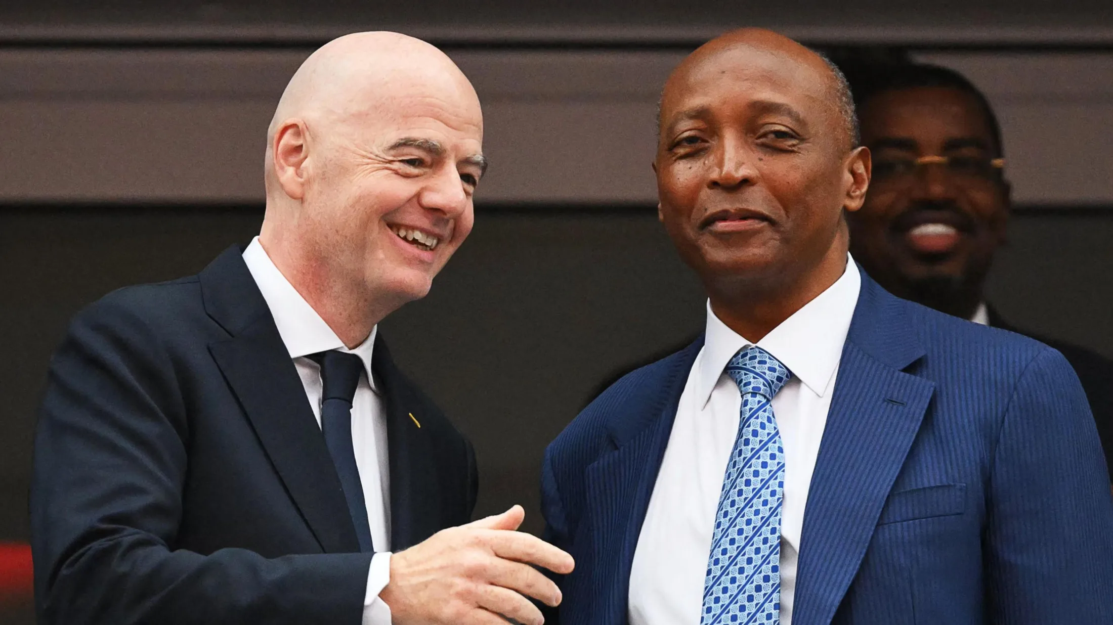 Fifa president Gianni Infantino, wearing a black jacket, white shirt and dark blue tie, smiles and holds up his right arm as he speaks to Caf president Patrice Motsepe, who is wearing a blue suit jacket, white shirt and blue patterned tie