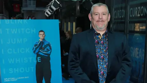 BBC upholds complaints over racial slur in Baftas broadcast Getty Images John Davidson stood in front of a film poster for I Swear