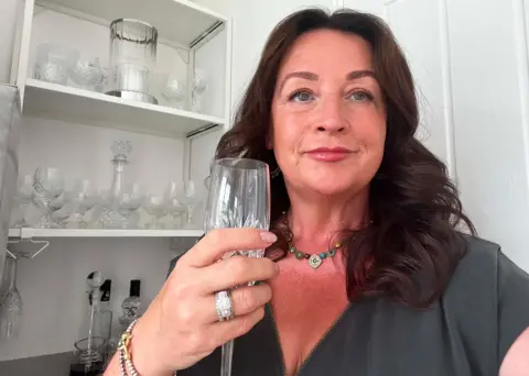 Ceri Evans Woman with long dark hair and thoughtful expression is holding a champagne glass. A large ring is visible on the fourth finger of her hand holding the glass.