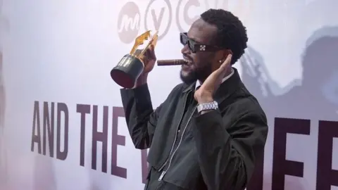 Dress made from 500 loaves stuns Africa Magic Viewers' Choice Awards BBC/ MOBOLAJI OLATUNDE Bucci Franklin, in a black outfit and wearing shades, is on the stage with his award.