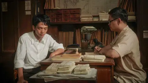 A Chinese box office hit sparks a debate about identity in Singapore Golden Village A man sits across another man at a desk. One of them is writing on a piece of paper.
