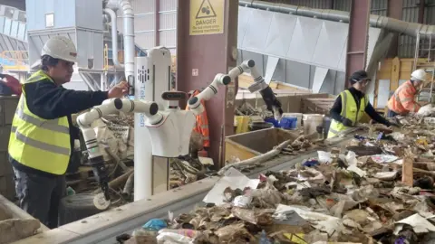 Alpha robot positioned at a conveyor belt carrying waste. Alpha has two very long arms and its upper body is attached to a column. 