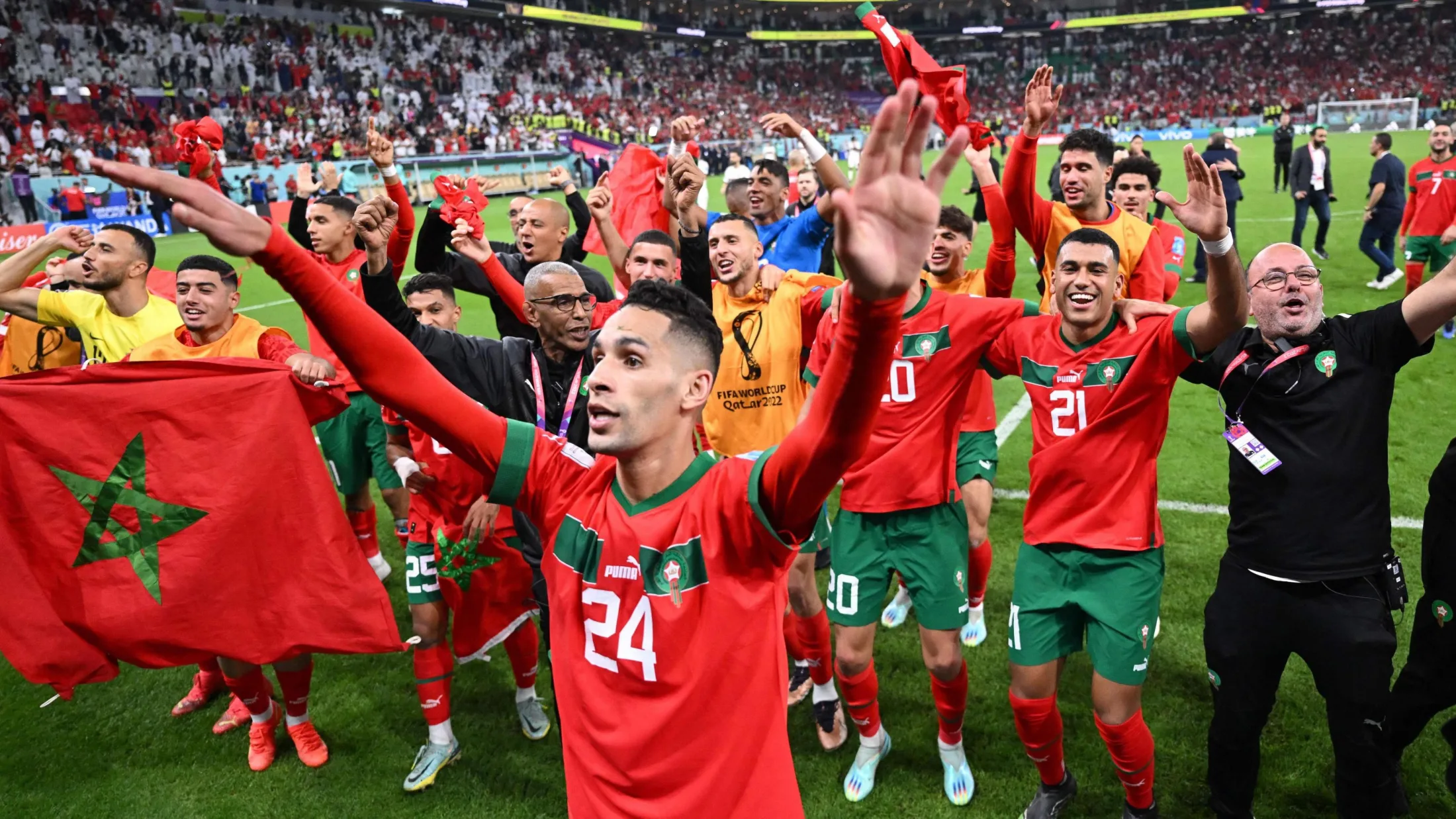 Morocco players and staff celebrate on the pitch after beating Portugal at the 2022 World Cup in Qatar. The group has organised itself into rough lines, placing arms around shoulders or waving arms in the air, with one player Badr Benoun, separated in the foreground close to the camera. Another player in an orange bib holds up a Morocco flag. The stands of an emptying stadium can be seen in the background