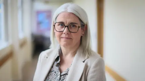 Secondary schools in Scotland cutting subjects due to teacher shortage 'crisis' Louise Moir standing in a school corridor. She has shoulder-length, grey hair and glasses, and is wearing a cream jacket and a patterned top.