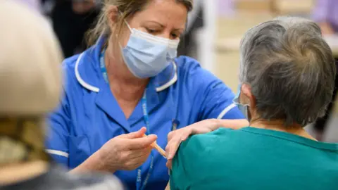 'World-first' vaccine designed by artificial intelligence Getty Images The image shows a healthcare setting where a medical professional is administering an injection to another person’s upper arm. The medical professional is wearing a blue uniform, a face mask, and holding a syringe carefully while preparing or delivering the shot. The seated individual has their sleeve slightly pulled up to expose the upper arm for the injection. The background appears softly blurred, with other people and elements suggesting a busy clinic or vaccination site.