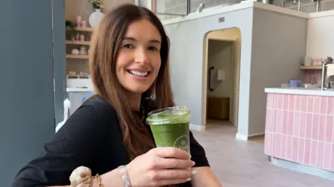 Why heatwaves hit women harder Jess Bloom Jess smiles, holding an iced matcha latte in a cafe. She has long brown hair and wears a black top