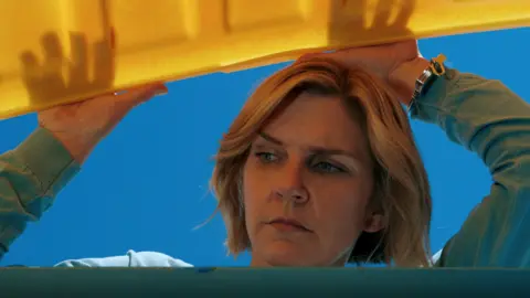 Apple TV Rhea Seehorn seen from below opening a large yellow lid of a large container and looking inside, in a still from Pluribus