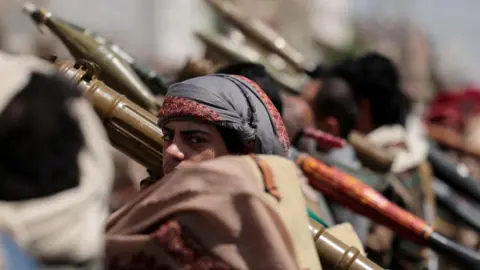 Reuters A man with a serious expression, wearing a head covering, holds what appears to be a bazooka over his shoulder, surrounded by other men holding the same weapons