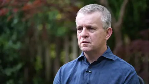 David Parfett speaking during an interview being conducted outdoors. He has short, grey hair and is wearing a blue shirt. Trees and leaves are in the background.