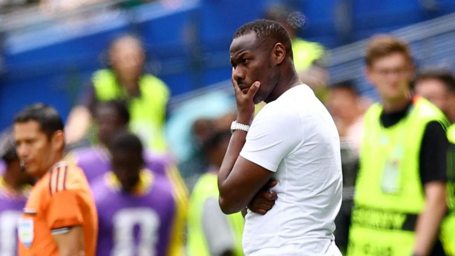 First Afcon, now World Cup - Senegal trapped in 'football hell' Pape Thiaw, visible from waist up wearing a white t-shirt, holds his left hand up to his chin in a thoughtful pose on the touchline during a football match. Out of focus in the background an assistant referee can be seen in an orange shirt, substitutes wear purple bibs and stewarding staff are in luminous yellow bibs