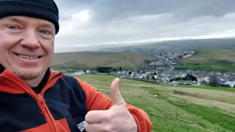 Instagram/Robert Charles Albon Robert Albon is dressed in an orange and black fleece. He is also wearing a black beanie hat. He is smiling and holding his thumb up. He is taking a selfie in front of a rural village surrounded by fields. 
