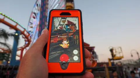 10 years of Pokémon Go and the millions still trying to catch 'em all PG/Bauer-Griffin A red and black phone showing the camera view of a location with an augmented Magikarp from Pokémon Go. In the blurred background there is a ferris wheel and a rollercoaster track.