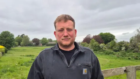 Rob Shield wearing a grey quarter zip sweatshirt standing against a fence with a grassy field behind him
