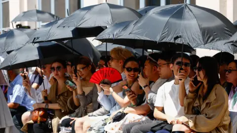 Reuters People shield themselves from the sun with umbrellas during Thom Browne’s Spring/Summer 2027 men’s collection show as part of Milan Fashion Week, in Milan, Italy, June 22, 2026.