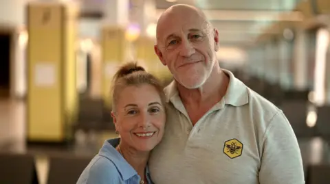 They became best friends – then discovered they were brother and sister Two adults standing close together in the foreground of an indoor waiting area. One person wears a light blue shirt over a white top and has hair tied back; the other wears a light grey polo shirt with a yellow badge featuring a bee symbol and has a tattooed forearm. Rows of dark chairs are visible behind them, with blurred columns, ceiling lights and windows extending along the corridor in the background. No visible bags, bins or markers are present.