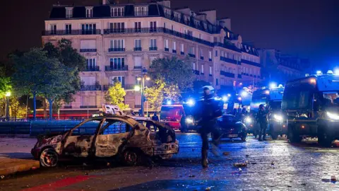 AFP via Getty Images A nighttime image of burnt out white car pictured infront of a row of French police vans with their blue lights flashing