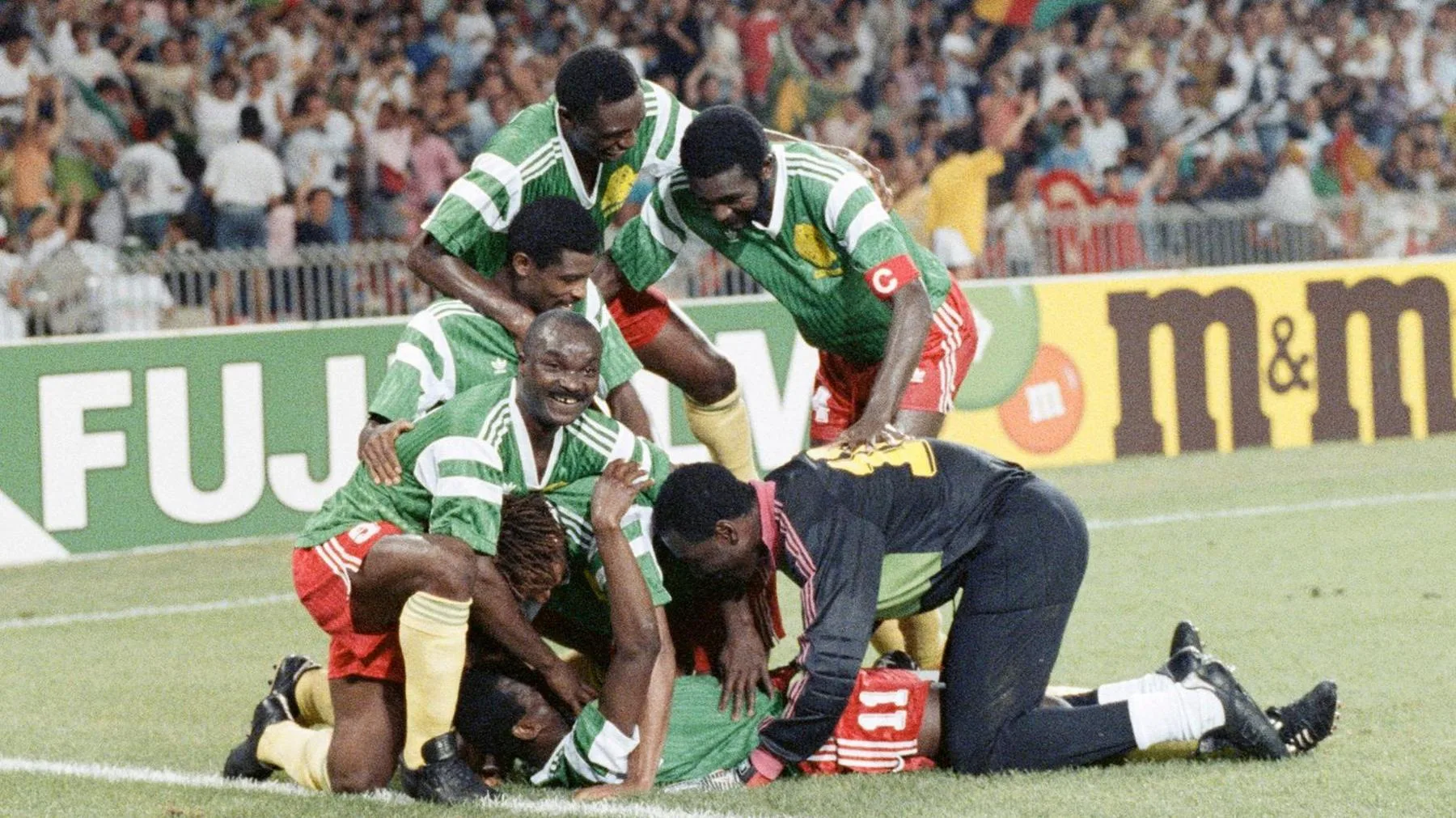 Cameroon outfield players in green shirts, red shorts and yellow socks, as well as the goalkeeper dressed mostly in black including his tracksuit bottoms, celebrate a goal at the 1990 World Cup by piling on top of Eugene Ekeke, who lies flat on his back as six players gather around him and offer congratulations. Advertising hoardings can be seen in the background, with a packed stand of fans out of focus in the distances. One player, Roger Milla, looks up from the pack and smiles towards the camera
