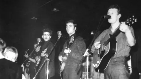 Getty Images George Harrison, John Lennon and Tony Sheridan play their guitars on stage in front of microphone stands to an audience in front of them.