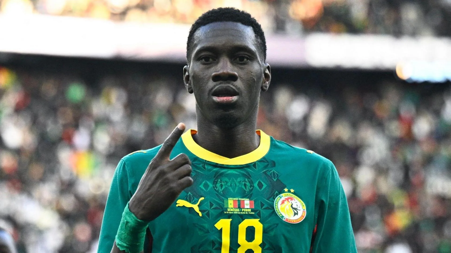 Ismaila Sarr, visible from chest up wearing a green Senegal shirt with the Senegalese and Peruvian flags on the chest alongside a yellow number 18, points to his own face with his right index finger as he looks with a serious expression towards the camera. A large crowd can be seen out of focus in the background
