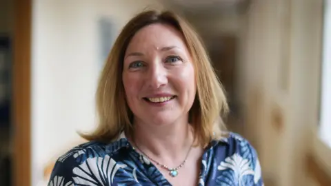 Secondary schools in Scotland cutting subjects due to teacher shortage 'crisis' Katrina Suèr standing in a school corridor. She has shoulder-length, brown hair and is wearing a blue and white patterned top.