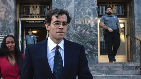 Getty Images Adam Mosseri, with dark curly hair and glasses, in a dark suit, white shirt, and spotted tie, seen against the marble-façade and steps of the Los Angeles County Superior Court.