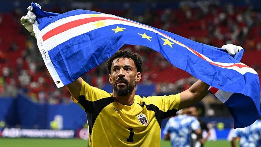 Cape Verde goalkeeper Vozinha, visible from chest up while wearing a yellow goalkeeping jersey with black trim, holds a billowing Cape Verde flag above his head with his arms outstretched. A half-empty stand at a football stadium can be seen out of focus in the background