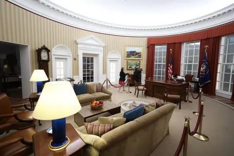 Talia Sprague/Bloomberg via Getty Images Stuffed chairs, lamps and dark wood furniture fill a replica of the Oval Office, with parts of the space sectioned off by red velvet ropes. Two people examine the space