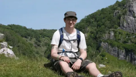 Callum Callum is wearing a white shirt and khaki shorts with a hat on. He is sat on a cliff with a rucksack on his back.