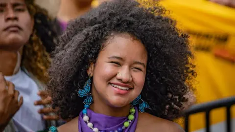 Catalina La Grande has dark curly hair. She is smiling in front of a crowd, and is wearing blue butterfly earrings.
