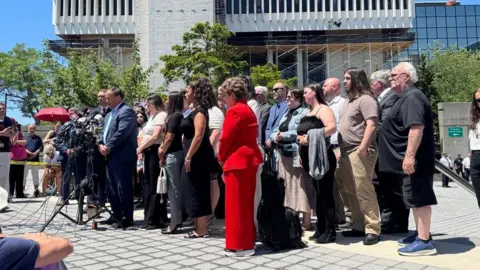 BBC News At a distance, a man stands at a mic with a group of people  surrounding him. they are in front oiif a courthouse outside with blue sky behind them