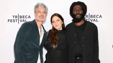 The notorious Cameroonian prison with its own record label Getty Images for Tribeca Festival Taika Waititi, Dione Roach and Steve Happi pose for a photo in front of a Tribeca Festival step and repeat