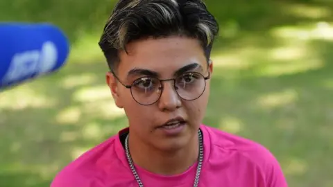 King Charles meets women's cricket team that is not allowed to exist Reuters Ekil Latifi, a woman with cropped dark hair with highlights, wearing black wire-framed glasses and a silver chain necklace with a bright pink T-shirt