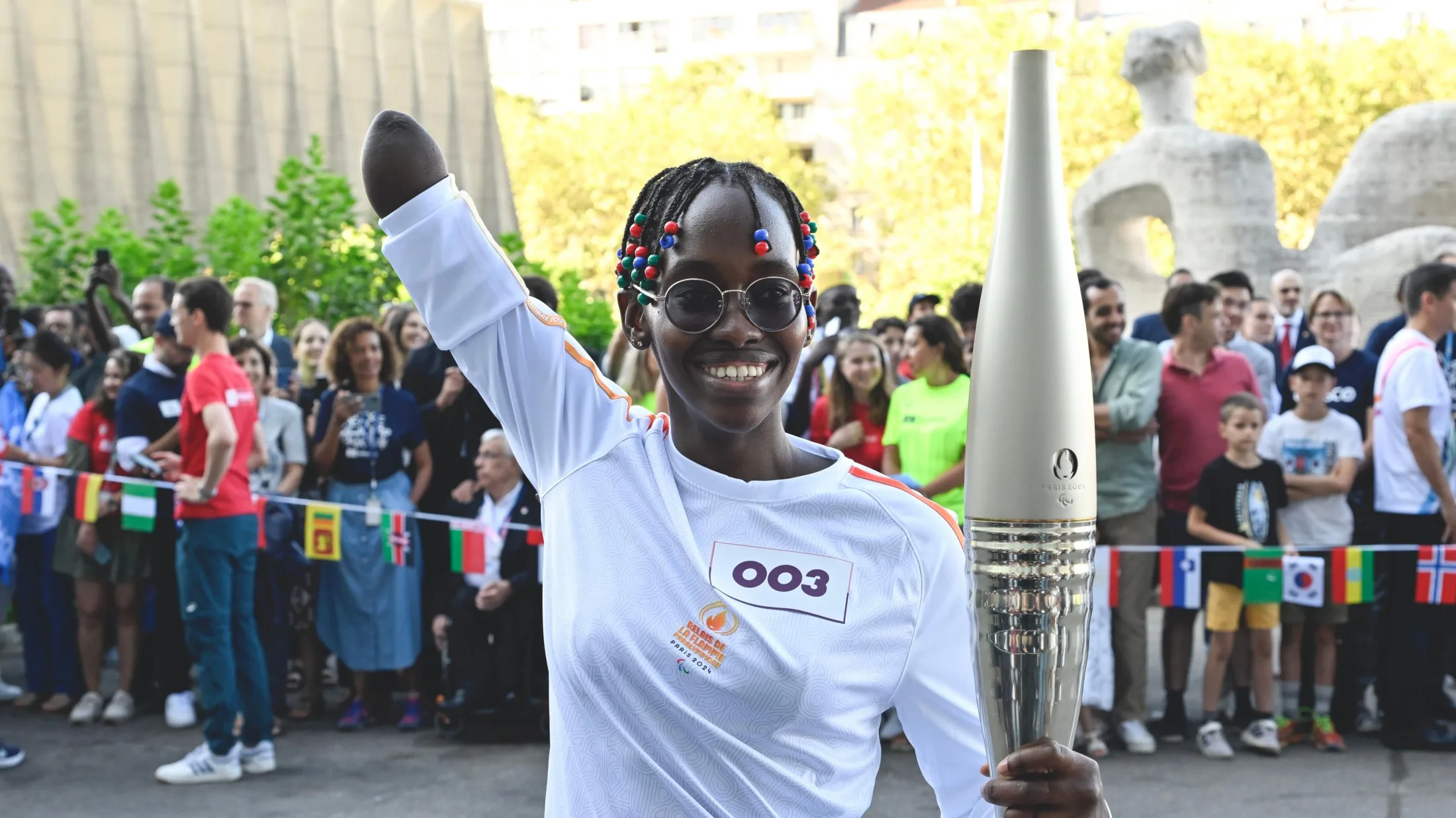 Husnah Kukundakwe, visible from waist up, smiles into the camera while taking part in the 2024 Paralympic torch relay. She wears a white sports top, round glasses and blue and red beads in her braided hair. In her left hand she holds the silver torch while she raises her right arm, which is missing its lower half, up in the air. Behind her stands a crowd of people being held back by a thin white tape from which hang the flags of various countries